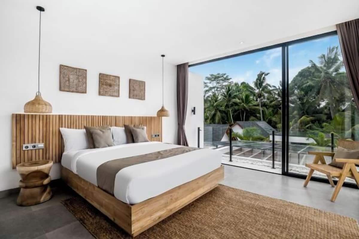 A modern bedroom with a large bed, wooden headboard, and a view of tropical vegetation through large glass doors.