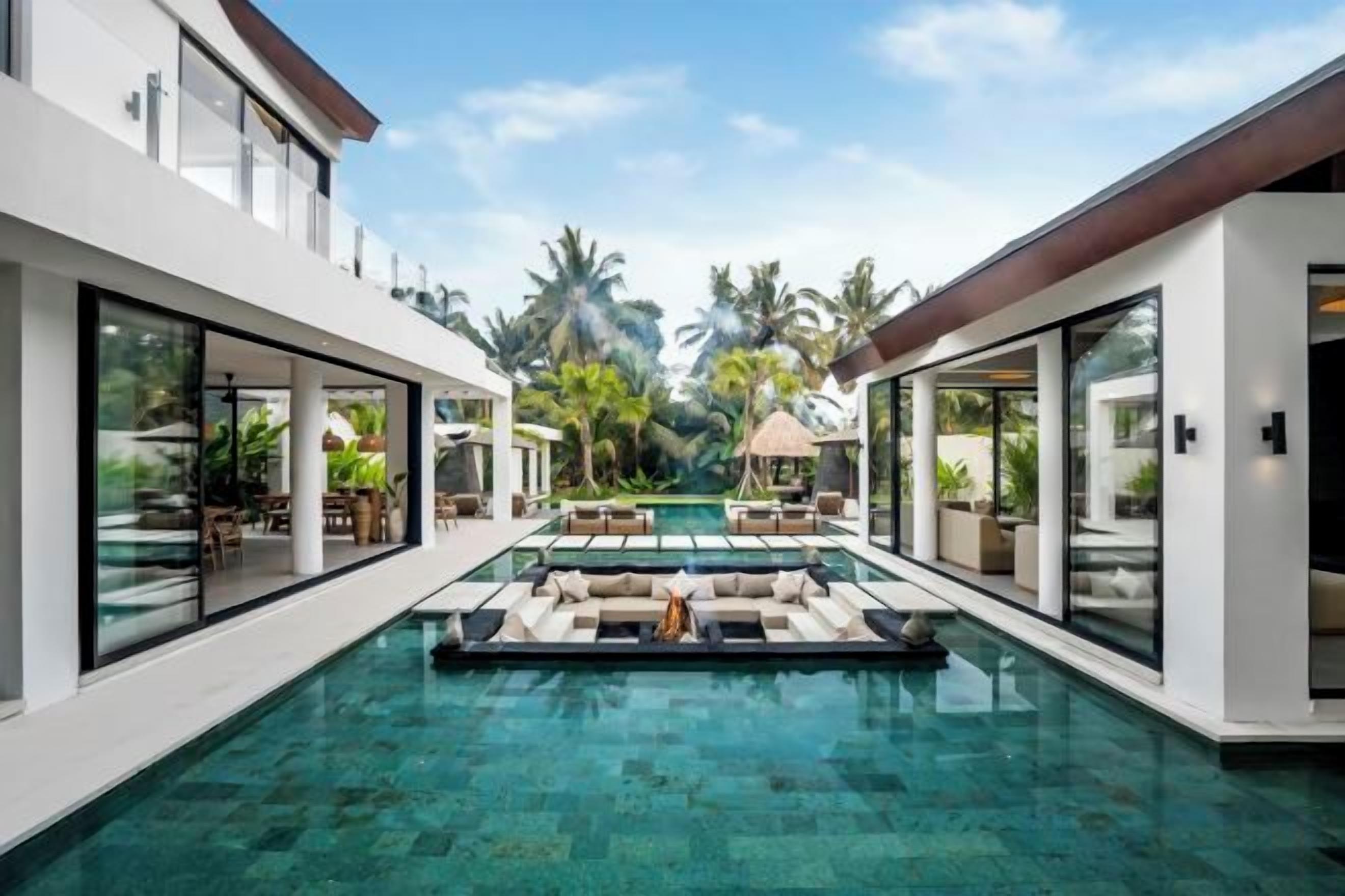 Un área de piscina moderna con paredes de vidrio, mobiliario exterior y vegetación tropical.