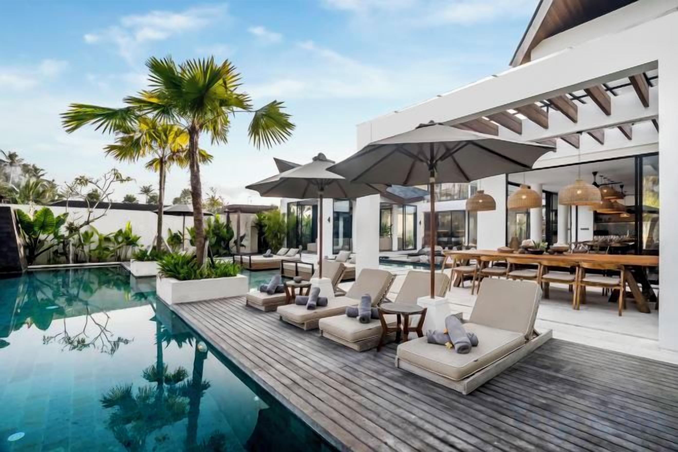 Un área junto a la piscina con sillones de descanso y sombrillas, una terraza de madera y un comedor con mesas y sillas.