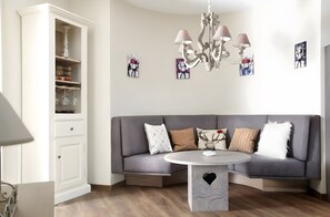 Un salon moderne avec un canapé d’angle gris, un meuble blanc contenant de la verrerie, une table ronde dont le pied est en forme de cœur et un lustre.