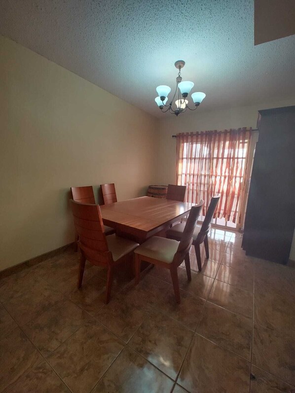 Une salle à manger avec une table et des chaises en bois, un lustre et une fenêtre avec des rideaux.