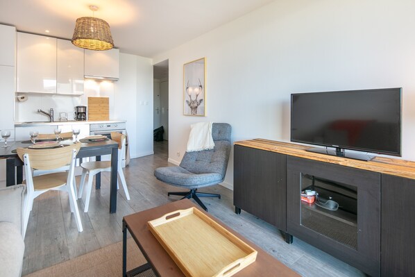Une cuisine moderne avec des meubles blancs, un coin repas avec une table et des chaises en bois, un fauteuil de bureau gris, un téléviseur posé sur un meuble en bois et un luminaire suspendu en osier.