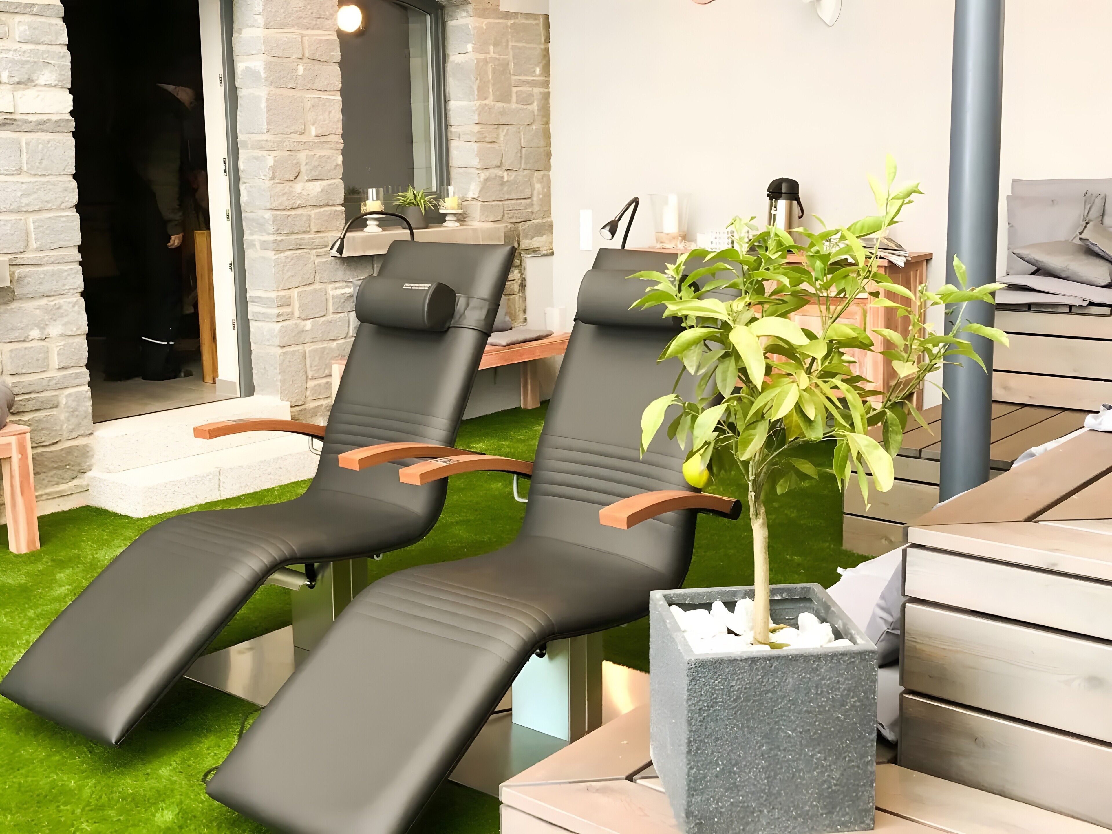 Two black lounge chairs with wooden armrests, a small potted plant, and a stone wall.