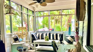 A sunroom with a white bench, striped cushions, a table, and a blue basket filled with items.