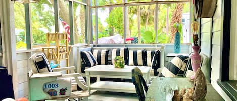 A sunroom with a white bench, striped cushions, a table, and a blue basket filled with items.