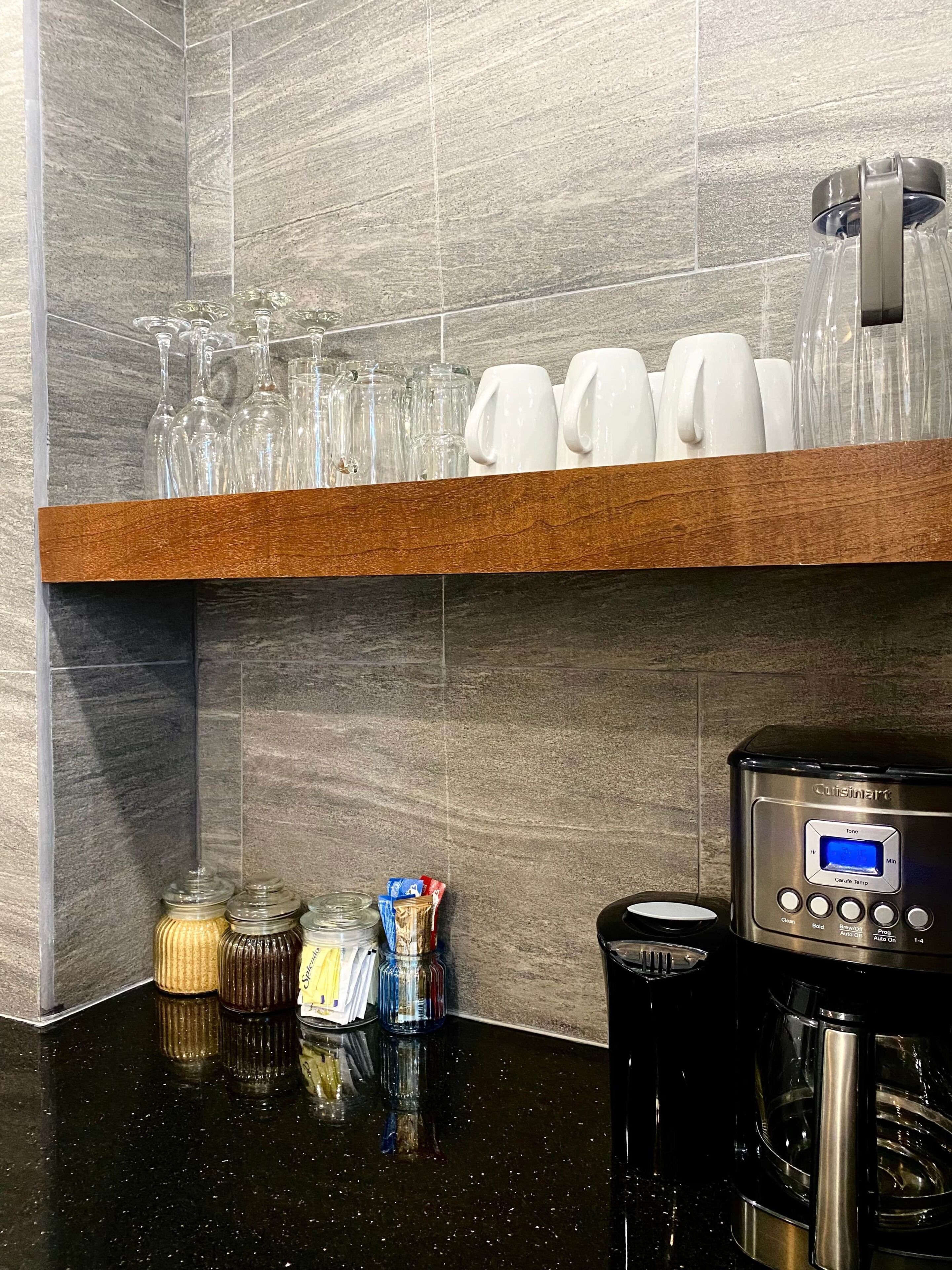 A kitchen area with a coffee maker, jars of condiments, and a shelf with glassware and white mugs.