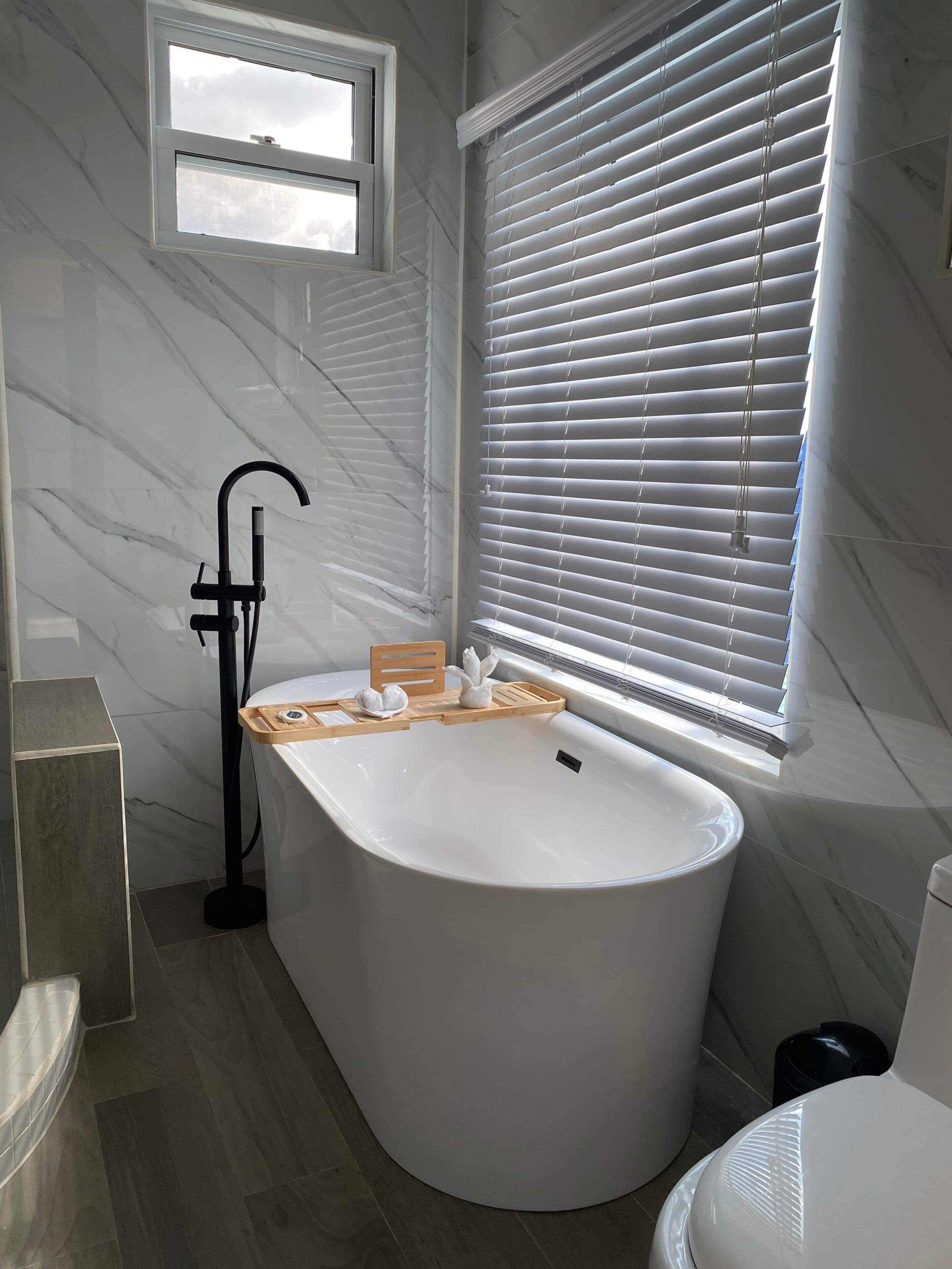 A modern bathroom with a freestanding bathtub, a window with blinds, and a marble wall.