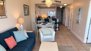 A living room with a dark blue sofa, a white coffee table, and a rug. There is a kitchen in the background with white cabinets and a dining area with white chairs. - Beach View ● Walks to Sand ● Pools & Hot Tub (Gulf Shores)