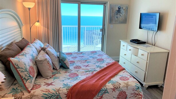 A bedroom with a bed, a dresser, a television, and a view of the ocean through a sliding door. - Beach View ● Walks to Sand ● Pools & Hot Tub (Gulf Shores)