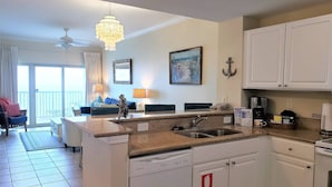 A modern kitchen with white cabinets, a granite countertop, and a dishwasher. There is a dining area with a table and chairs, a chandelier, and a painting on the wall. - Beach View ● Walks to Sand ● Pools & Hot Tub (Gulf Shores)