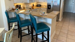 A kitchen with white cabinets, a stainless steel refrigerator, a microwave, and a coffee maker. There are four blue bar stools and a small island with two cups on it. - Beach View ● Walks to Sand ● Pools & Hot Tub (Gulf Shores)