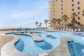 A swimming pool with a curved design, surrounded by lounge chairs and palm trees, with a building in the background. - Gulf View Condo · Hot Tub · Sauna · Indoor Pool (Gulf Shores)