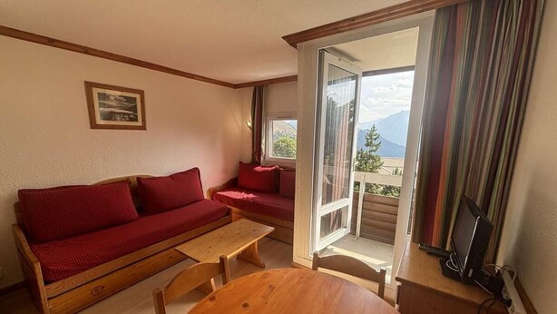 A cozy room with a wooden sofa, a small table, and a view of mountains through a sliding door. - 300m from ski lifts, 1st floor, balcony, tv, ski locker, 28m², Alpe d'Huez (L'Alpe d'Huez)