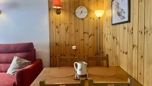 A cozy room with a wooden wall, a red sofa, a wooden dining table with chairs, a white clock, and a framed picture.