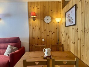 A cozy room with a wooden wall, a red sofa, a wooden dining table with chairs, a white clock, and a framed picture.