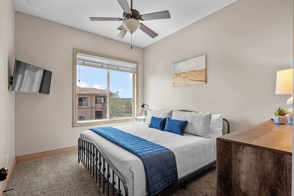 A bedroom with a bed, a ceiling fan, a window, a TV, and a painting on the wall. - The Overlook at Coral Springs (Hurricane)
