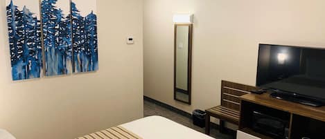 Ett hotellrum med en säng, en platt-TV, en tavla med ett trädmotiv, en spegel och ett litet bord med en mikrovågsugn.