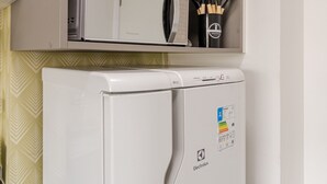 A kitchen with a white refrigerator, a microwave, and a shelf above containing a vase with flowers and kitchen utensils.