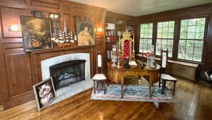 A room with a fireplace, wooden paneling, a dining table with chairs, and a large window with a view of greenery. - The House Hotels - Acadia Farms (Northfield)