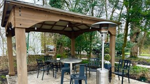 A covered outdoor seating area with tables and chairs, a central heating unit, and a pergola structure. - The House Hotels - Acadia Farms (Northfield)