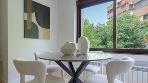 Un comedor con una mesa redonda y seis sillas, un cuadro abstracto moderno en la pared, y vistas a árboles y edificios a través de un ventanal grande.