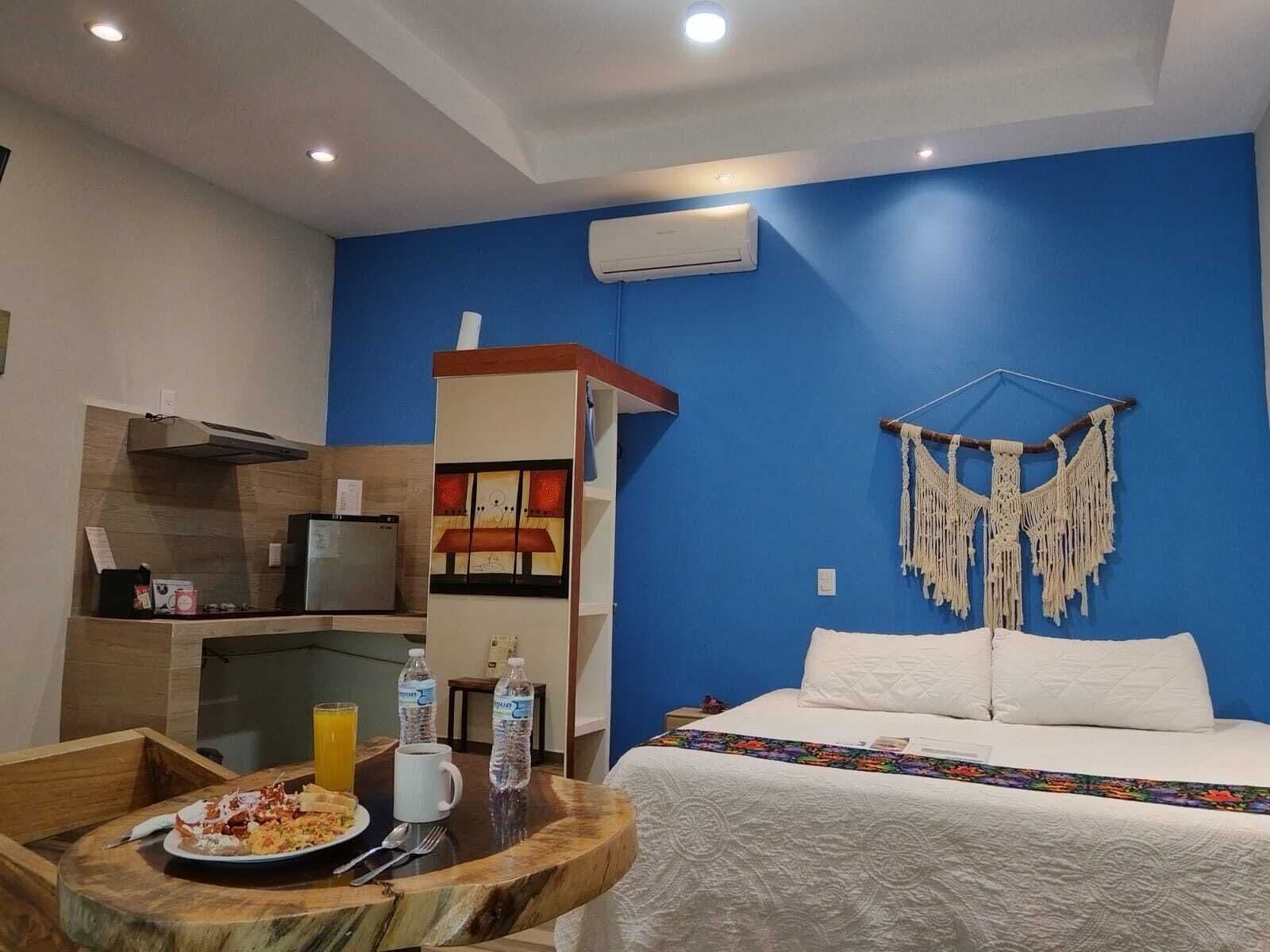 Habitación de hotel con cama, una mesa de comedor con desayuno, kitchenette y un tapiz de macramé en la pared.