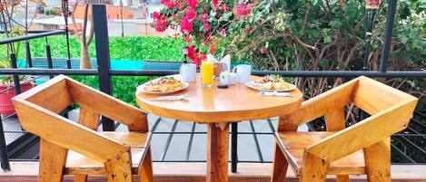 Una mesa de madera con dos sillas, un vaso de jugo de naranja y platos de comida en un balcón con vistas a edificios y flores rosadas.