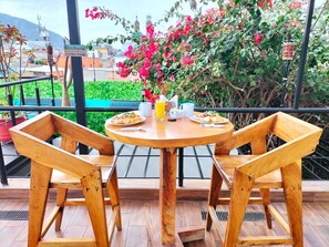 A wooden table with two chairs, a glass of orange juice, and plates of food on a balcony with a view of buildings and pink flowers.