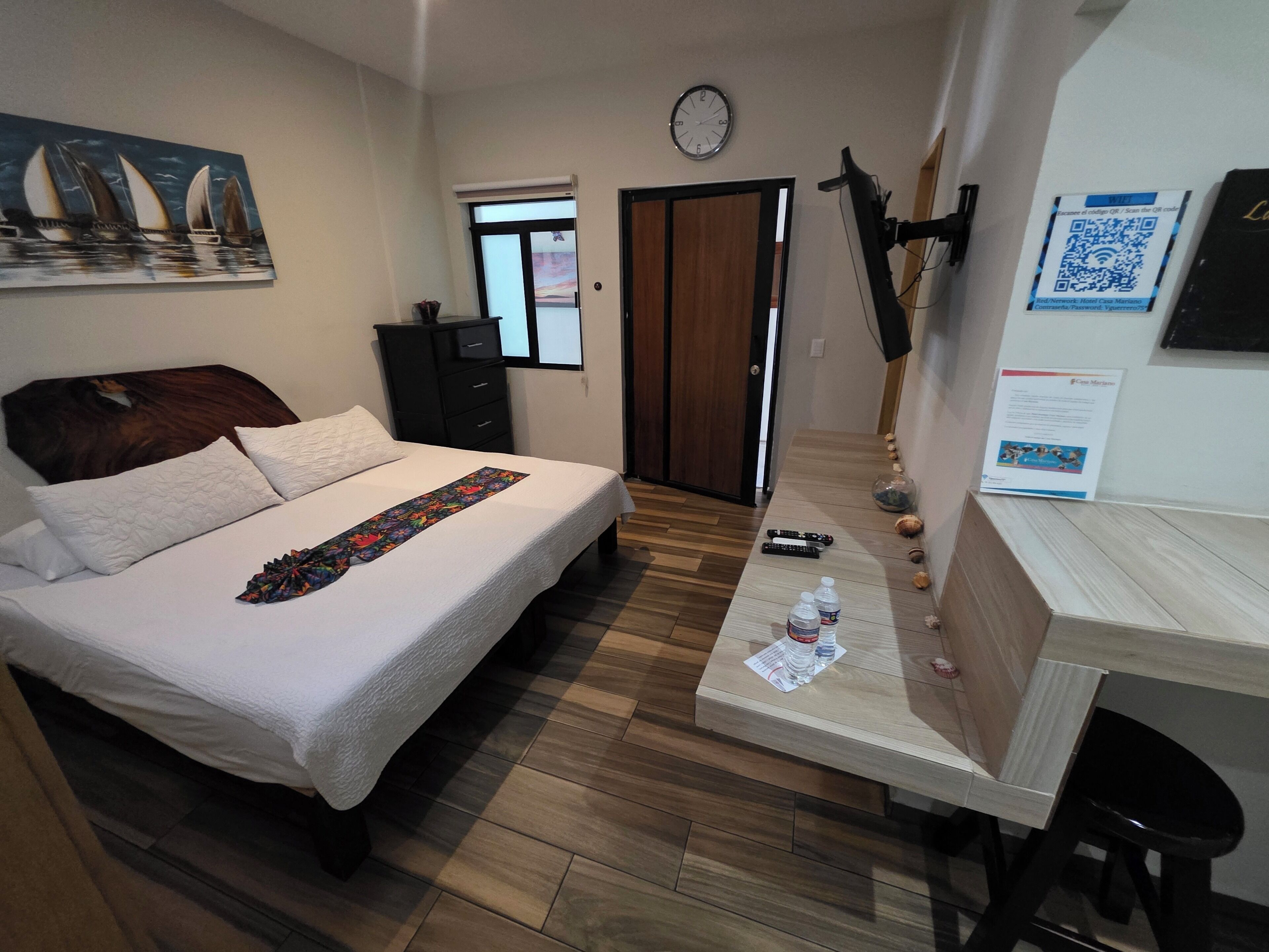 A hotel room with a bed, a desk, a clock, and a painting on the wall.