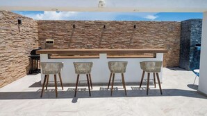 Ein Außenbereich mit Bar, der von einer Steinwand gesäumt wird, einer Holztresore und drei Korbstühlen.