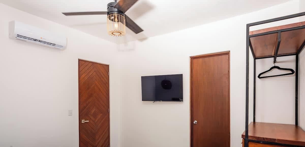 A room with two wooden doors, a ceiling fan, an air conditioning unit, a mounted television, and a metal shelf with a hanger.