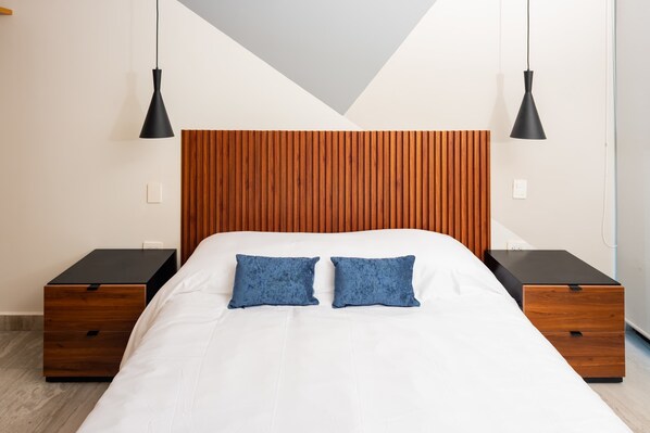 Un dormitorio moderno con cabecera de madera, dos mesitas de noche, cama con ropa blanca y dos almohadas azules.