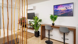 A modern living room with a sofa, a wooden desk, two bar stools, a TV, and a large window with a view of birds flying. - Charming Stay – 5 Minutes from Walmart & 5th Avenue (Playa del Carmen)