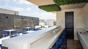 A modern outdoor bar area with a long counter, blue chairs, and a stone wall with a logo. - Charming Stay – 5 Minutes from Walmart & 5th Avenue (Playa del Carmen)