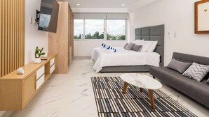 A modern bedroom with a bed, a sofa, a TV mounted on the wall, and a window with a city view. - Warmth and Charm in Harmony (Cancún)