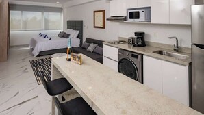 A modern kitchen with a microwave, coffee maker, and washing machine. There is a bed with a gray headboard and a gray sofa. - Warmth and Charm in Harmony (Cancún)