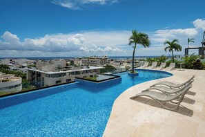 Une piscine entourée de chaises longues et de palmiers, avec des bâtiments modernes en arrière-plan et un ciel dégagé.