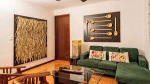 Un salón con un sofá verde, sillas de madera, una mesa de centro de vidrio y una obra de arte en la pared.