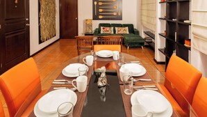 Un comedor con una larga mesa dispuesta para seis personas, rodeada de sillas color naranja, con un sofá verde y una estantería al fondo.