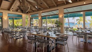 A spacious outdoor dining area with multiple tables and chairs, large windows offering views of the ocean and palm trees, and a wooden ceiling with hanging lights. - Studio Double@ Elysian Beach Resort (St Thomas)