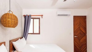 Un dormitorio con cama, lámpara de mimbre colgante, ventana con cortinas, puerta de madera y un equipo de aire acondicionado.