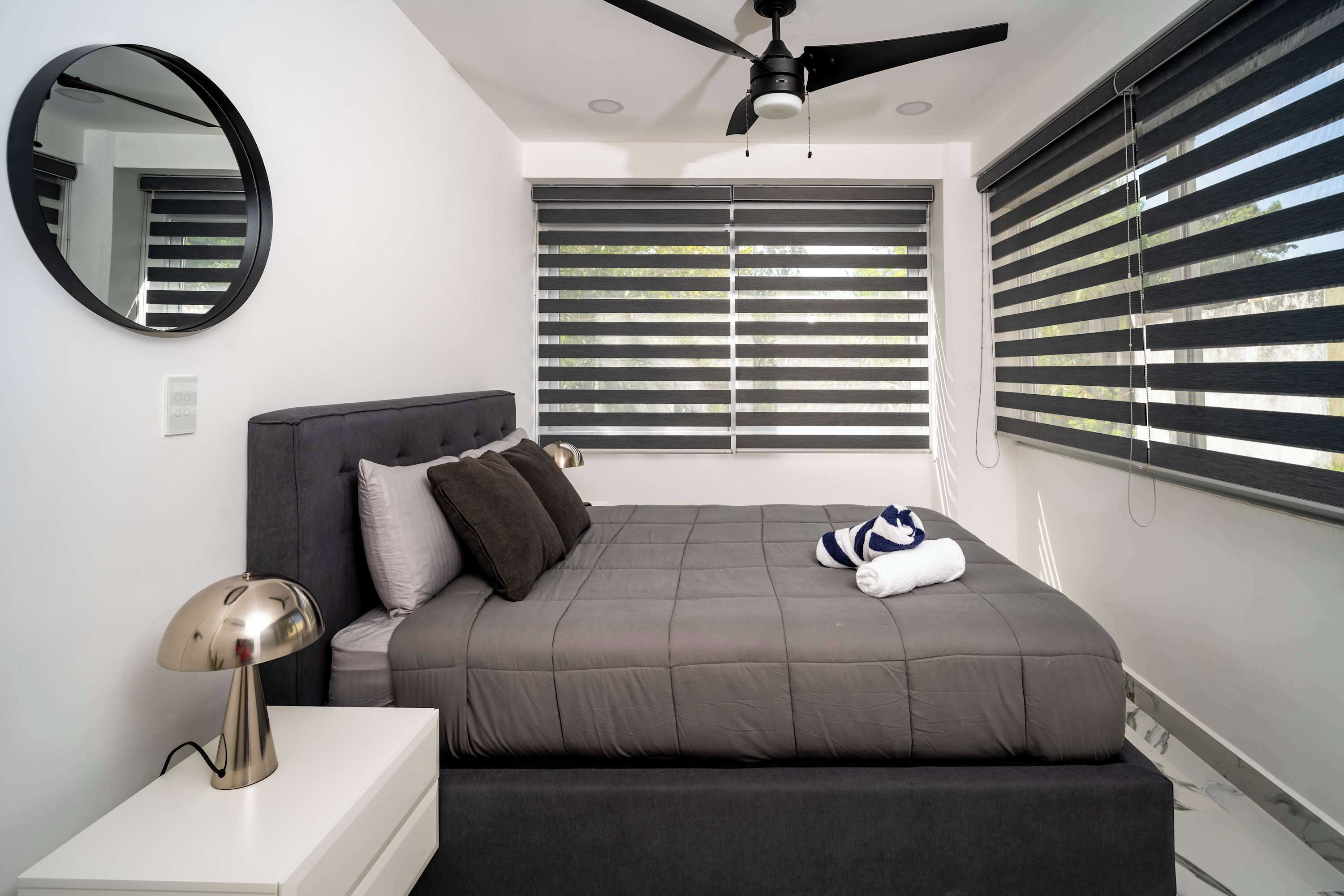 Un dormitorio moderno con cama, mesita de noche, lámpara, ventilador de techo y ventanas grandes con persianas.