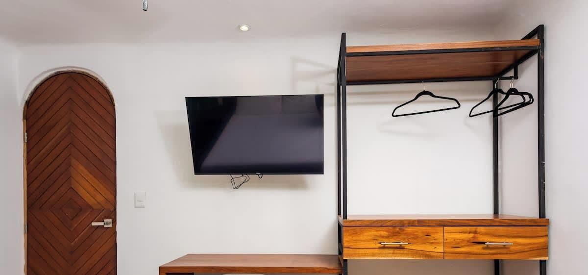 A room with a wooden door, a television mounted on the wall, a wooden cabinet with drawers, and a metal shelf with hangers.