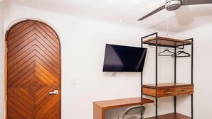 Ein Zimmer mit einer Holztür im Fischgrätmuster, einem an der Wand befestigten Fernseher, einem Metallbettgestell und einem Holzbüro sowie einem Deckenventilator.