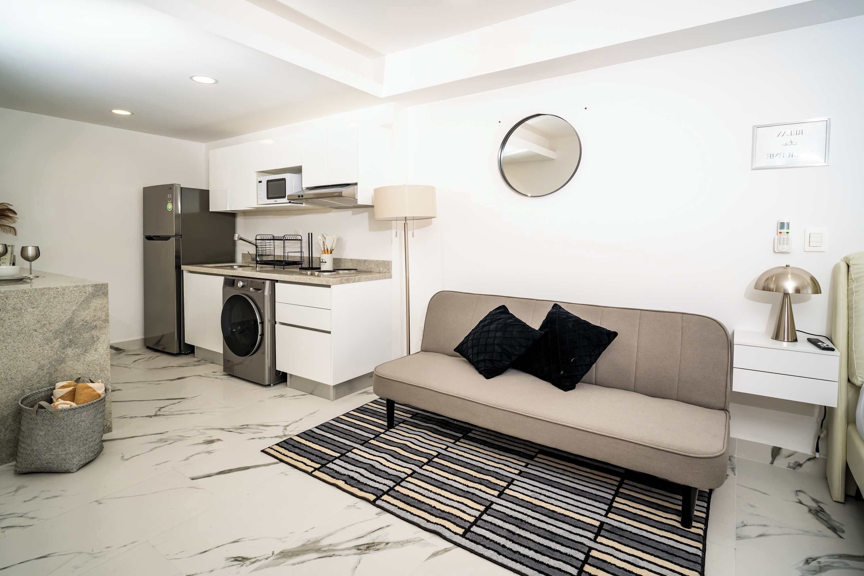 Um apartamento moderno com cozinha compacta, sofá, espelho e mesa de cabeceira com abajur.