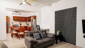 A living room with a sofa, a dining table with chairs, a kitchen area with wooden cabinets, and a black roller blind. - Villa for groups in excellent location (Playa del Carmen)