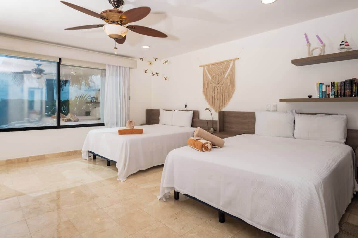 A hotel room with two beds, a ceiling fan, a large window with curtains, a macrame wall hanging, and a shelf with books and decorative items.