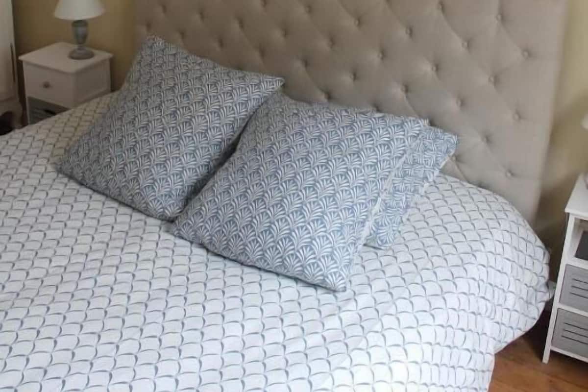 Een slaapkamer met een gestoffeerd hoofdeinde, beddengoed met een patroon, een nachtkastje met een lamp en een wanddecoratie waarop een toren met de tekst 'SUD' staat afgebeeld.