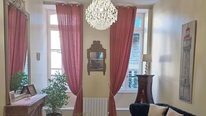 Un salon avec un lustre, des rideaux rouges, un canapé noir, une petite table et une plante en pot.