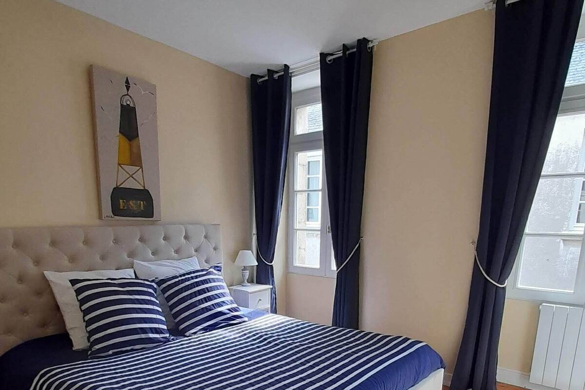 Een slaapkamer met een groot bed, een gestuwd hoofdeinde, een nachtkastje, een lamp, een schilderij en een raam met gordijnen.